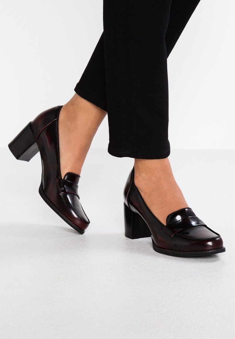 Mocassins en cuir verni bordeaux avec un talon épais, présentant un accent cousu et un bout arrondi. Portés avec un pantalon noir sur un sol blanc.
