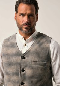 Chaleco gris y beige con una textura suave, con escote en V, cinco botones y un bolsillo en el pecho. Llevado sobre una camisa blanca de cuello.