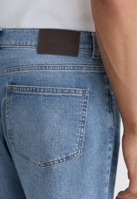 Lähikuva vaaleansinisten farkkujen takataskusta ja vyötärönauhasta, jossa on tummanruskea Reiss-merkkilappu, käytetty valkoisen ribbineulotun topin kanssa.