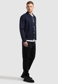 Homme portant une veste marine, une chemise blanche, un pantalon noir et des chaussures noires, debout et regardant sur le côté sur un fond clair uni.