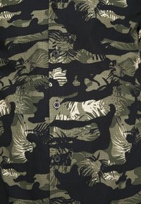 Groen camouflage shirt met een mix van donkere en lichte groentinten, een gestructureerd patroon van bladeren en diverse vormen, ontwerp met knoopsluiting aan de voorkant.