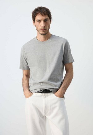 Mann mit kurzen lockigen Haaren, der ein graues kurzärmeliges T-Shirt und weiße Hosen trägt und mit den Händen in den Taschen vor einem einfarbigen Hintergrund steht.