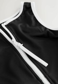 Sort beklædningsgenstand med en glat, strækbar tekstur, udstyret med en hvid lynlås og kant langs halsudskæringen. Lynlåstrækker er af hvid plastik med mærkning.