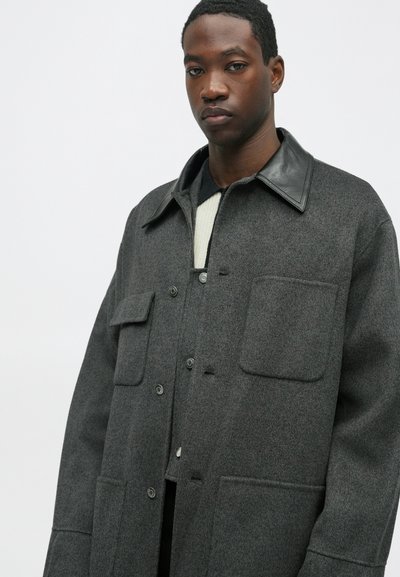 Vīrietis ģērbjas tumši pelēkā vilnas mētā ar lielām priekšējām kabatām un melnu ādas apkakli, virs viegla džempera, stāvot uz vienkārša fona.