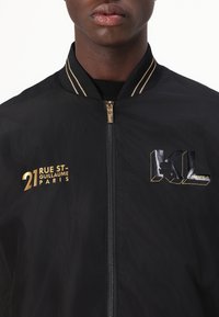 Black bomber jacket made of lightweight material, featuring gold hardware, striped collar, text "21 RUE ST-GUILLAUME PARIS," and "KL LAGERFELD" logo.