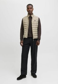 Beige gesteppte Weste mit hohem Kragen, getragen über einem dunkelbraunen Strickpullover. Schwarze Hose und Schuhe runden das Outfit ab. Einfaches Design, stilvolle Hardware.