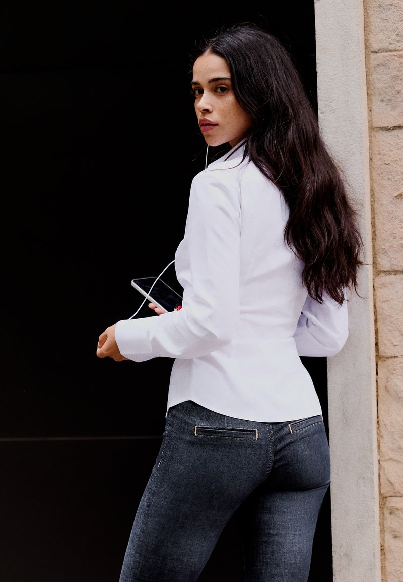 Jeune femme aux longs cheveux foncés portant une chemise blanche et un jean foncé, tenant un smartphone avec des écouteurs, regardant par-dessus son épaule.