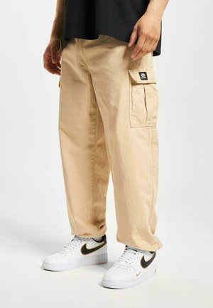 Pantaloni cargo beige con una vestibilità rilassata, dotati di due tasche laterali e una toppa con etichetta. Abbinati a sneaker bianche con dettagli neri.