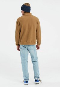 Braune Fleecejacke, blaue Strickmütze, hellblaue Jeans und mehrfarbige Sneakers. Lässiger Schnitt mit strukturierten Materialien und kontrastierenden Farben.
