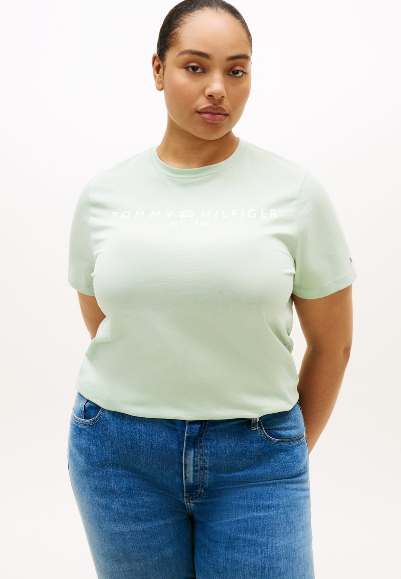 Ανοιχτό πράσινο βαμβακερό t-shirt με κοντά μανίκια, με το λογότυπο "TOMMY HILFIGER EST. 1985" τυπωμένο με λευκά γράμματα στο στήθος. Ορατό το τζιν παντελόνι.