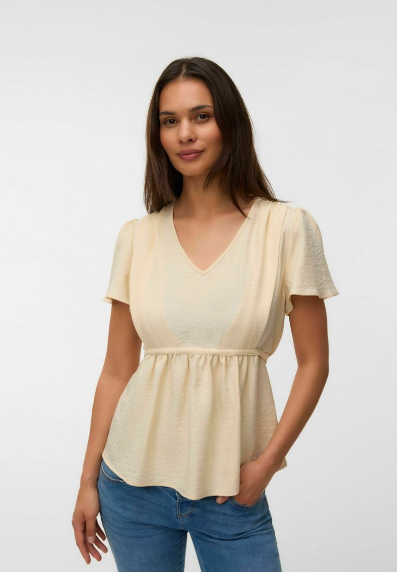 Femme aux longs cheveux foncés portant un chemisier crème à manches courtes et col en V, et un jean bleu, debout avec une main dans la poche sur fond blanc.