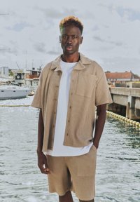Homme aux cheveux courts et bouclés portant une chemise beige texturée à manches courtes et un short avec un t-shirt blanc, debout près du quai avec des bateaux et des bâtiments.