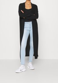 Cárdigan largo negro superpuesto sobre un top ajustado negro, combinado con vaqueros pitillo azul claro y zapatillas blancas. Textura suave, diseño de frente abierto.