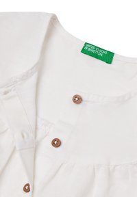 Top de algodón blanco con escote redondeado, botones de madera y dobladillo con volantes. Etiqueta verde con el logo "United Colors of Benetton" visible.