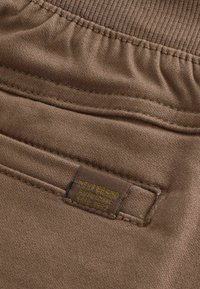 Brązowy materiał o gładkiej teksturze, z prążkowanym paskiem w talii i metką z napisem "NEXT BRAND AUTHENTIC EST. 1982" w kolorze żółtym.