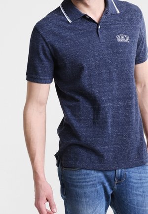 Mężczyzna w granatowej koszulce polo z krótkim rękawem z białymi wykończeniami i logo "GAP", w połączeniu z niebieskimi dżinsami, na jednolitym tle.