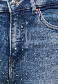 Šviesiai mėlynos spalvos džinsai iš džinso audinio su išblukusia faktūra, išsklaidytomis skaidriomis akutėmis ir sidabrine sagos užsegimu juosmens srityje.