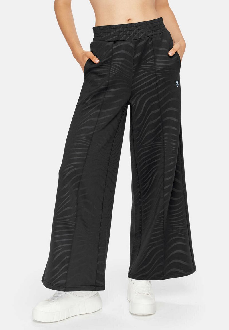 Schwarze weit geschnittene Hose aus glattem Stoff mit einem dezenten Wellenmuster, ausgestattet mit einem elastischen Bund und seitlichen Taschen. Kombiniert mit weißen Turnschuhen.