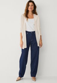 Cardigan lungo beige chiaro sopra un top bianco, abbinato a jeans a gamba larga blu scuro a vita alta e scarpe con tacco nudo.