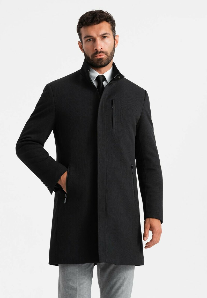 Cappotto di lana nero con colletto alto, maniche lunghe, tasca con zip sul petto e tasche laterali. Indossato sopra una camicia bianca e una cravatta.