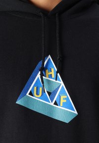 Schwarze Kapuzenjacke mit einem geometrischen Dreiecksdesign in Blautönen, verziert mit gelben Buchstaben "UHF" und aus Baumwollmaterial gefertigt.