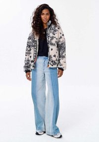 Mikina z flísu s čiernym kvetinovým vzorom na krémovom pozadí, plný zips a elastické manžety, skombinovaná s voľnými svetlo modrými džínsami a teniskami.