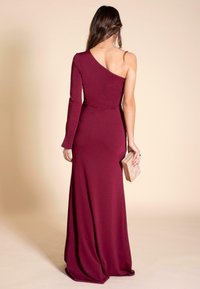 Vestido burdeos de un solo hombro con manga larga, diseño ajustado, largo hasta el suelo y textura suave, sostenido con un pequeño bolso geométrico.
