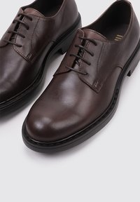 Chaussures en cuir brun avec un bout arrondi, un laçage et des coutures contrastées. Présentent une texture lisse et une semelle en caoutchouc noire.