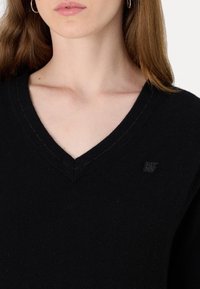 Pull noir en v en tissu tricoté, avec un détail de logo subtil. La texture est lisse, avec des coutures distinctes le long des bords.