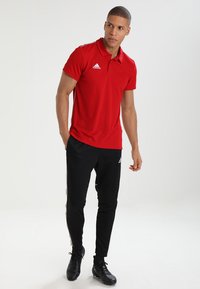 Rotes Polohemd mit Kragen, das ein Logo auf der Brust trägt. Kombiniert mit schwarzen schmal geschnittenen Trainingshosen mit weißen seitlichen Streifen.