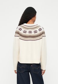 Cremefarvet ribstrikket sweater med en mønstret bærestykke med geometriske designs i brun og beige. Cropped længde og afslappet pasform.
