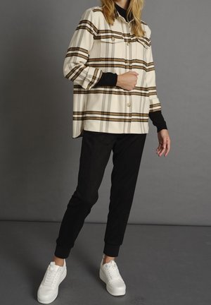 Chemise surdimensionnée à carreaux blancs et bruns avec deux poches sur la poitrine, associée à un pantalon noir fuselé et des baskets blanches. Convient pour une tenue décontractée.