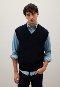 Gilet lavorato a maglia blu navy con scollo a V, indossato sopra una camicia di jeans azzurri. Presenta maniche arrotolate e jeans di denim scuro.