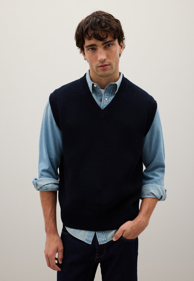Gilet lavorato a maglia blu navy con scollo a V, indossato sopra una camicia di jeans azzurri. Presenta maniche arrotolate e jeans di denim scuro.