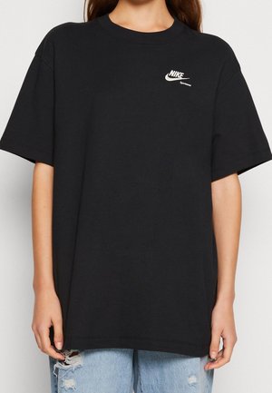 Femme portant un t-shirt noir ample Nike Sportswear et un jean bleu clair déchiré, debout, les bras détendus le long du corps.