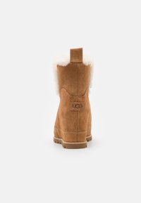 Botte de cheville en daim marron avec un col en fausse fourrure blanche et duveteux, offrant une semelle texturée et un petit patch logo à l'arrière.
