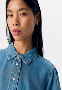 Heledsinine denim särk terava kraega, millel on pärlitega nööbid ja lõtv lääne. Kangas on pehme, veidi tekstureeritud välimusega.