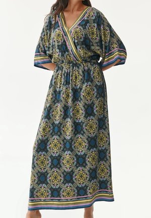 Kobieta w długiej sukience w wzory w niebieskie, zielone i żółte geometryczne wzory oraz paski na rękawach i dole sukienki.