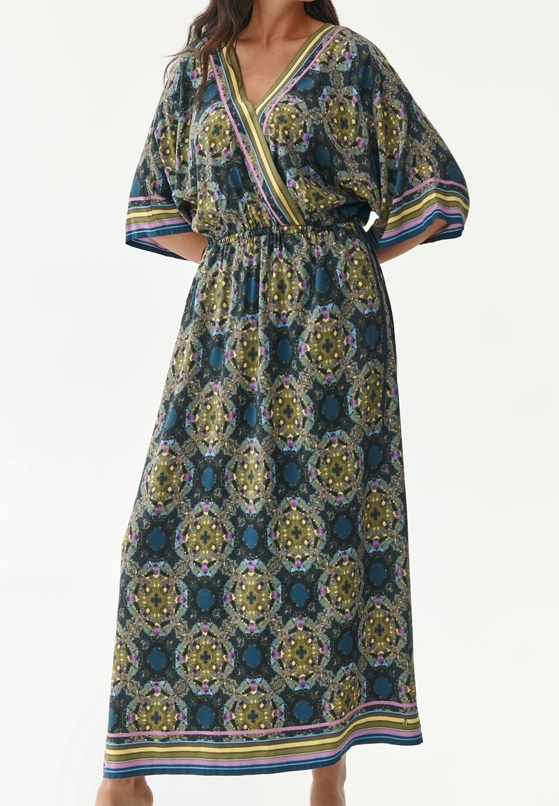 Femme portant une longue robe à motifs avec des dessins géométriques bleu, vert et jaune et une bordure rayée aux manches et à l'ourlet.