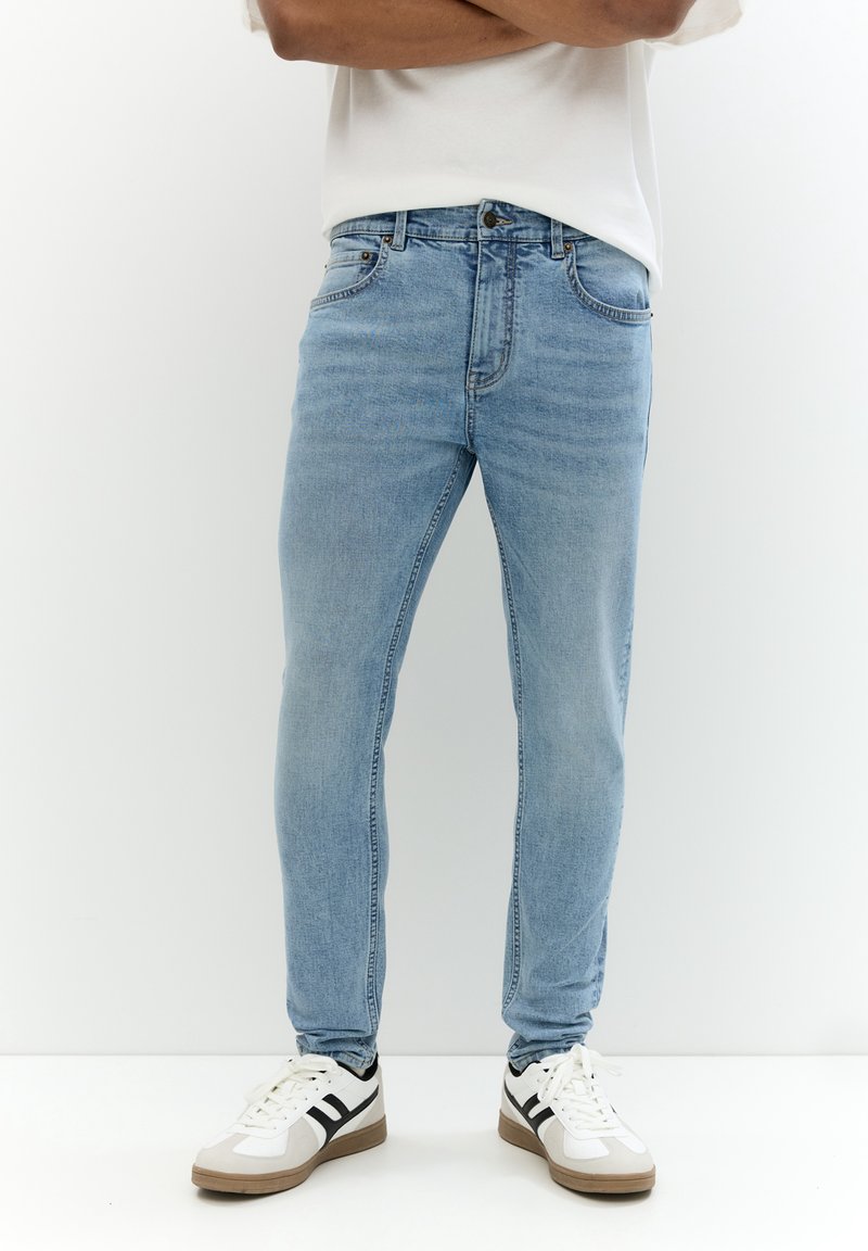 Homme portant un jean slim bleu clair et des baskets blanches avec des bandes noires, debout devant un fond blanc uni.