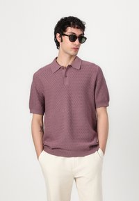 Short-sleeve polo shirt in dusty rose with a textured zigzag pattern, two-button placket, and ribbed collar. Paired with light-colored trousers.