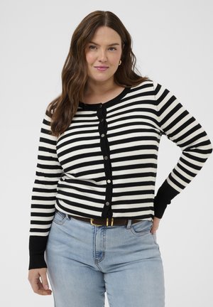 Kvinde iført en sort og hvid vandretstribet knaptrøje, lyseblå jeans og et brunt bælte, stående med den ene hånd på hoften.