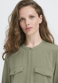 Jeune femme aux longs cheveux ondulés portant une chemise boutonnière vert olive clair avec des poches poitrine, regardant calmement l'objectif sur un fond uni.