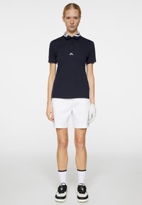 Marinblå polo med randig krage, vit logotyp, matchad med vita shorts. Bärs med en golfhandske, svart och vita sneakers, samt vita strumpor.