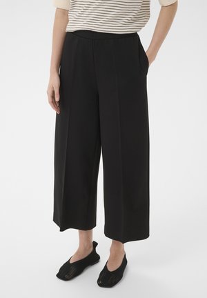 Nő fekete, bőszárú, rövidített nadrágot, csíkos rövid ujjú felsőt és fekete hálós lapos cipőt visel, fehér háttéren áll.