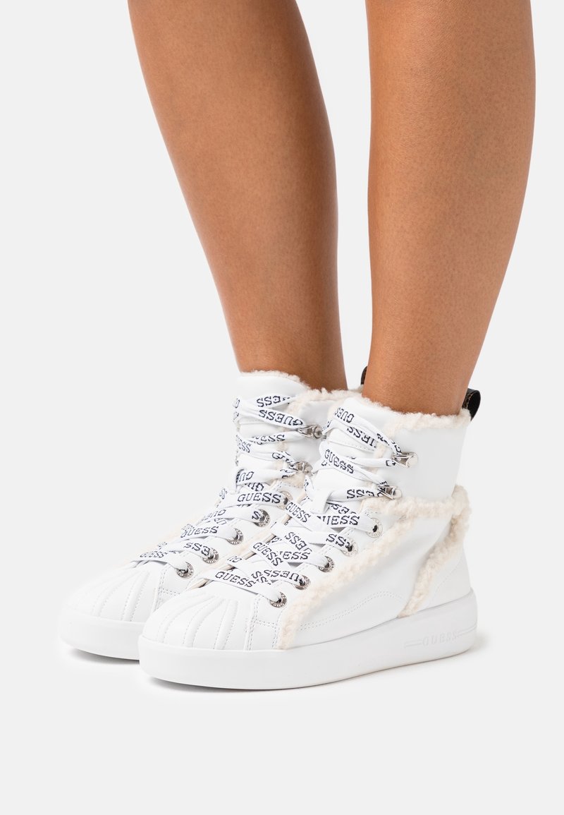 Baskets montantes blanches avec doublure en polaire et lacets griffés "GUESS" portées aux pieds d'une personne sur un fond uni.