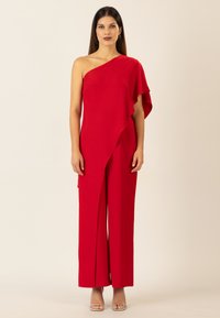 Jumpsuit rosso a spalla unica con drappeggio fluente, gamba ampia, tessuto liscio e un accento a strati sulla spalla. Sandali con tacco argento sotto.