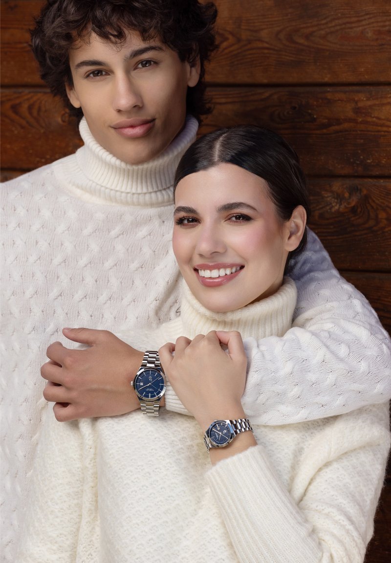 Due persone indossano maglioni bianchi a collo alto in maglia grossa. Entrambi mostrano orologi con quadrante blu, bracciali in acciaio inossidabile e dettagli in argento.