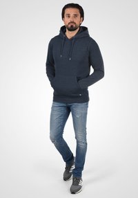 Dunkelblaues Hoodie mit einer Kordelzugkapuze, vorderen Taschen und langen Ärmeln; kombiniert mit hellblauen, distressed Jeans und grauen Sportschuhen.