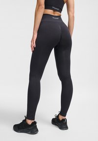 Svarta leggings gjorda av texturerat, elastiskt material; hög midja med logoprenom närmast midjebandet; ihopparat med svarta träningsskor.